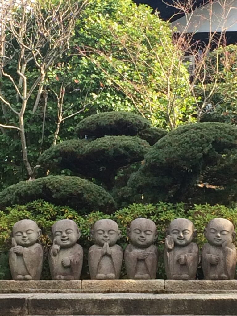 京都の寺の境内、緑の植え込みの前に並ぶ六体の小さなお地蔵様、合掌や笑顔の表情でそれぞれ佇む
