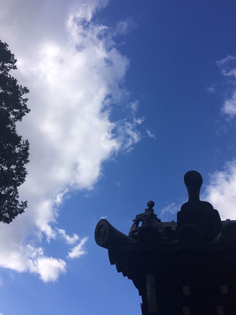 青空と白い雲を背に、寺院の屋根の軒先が黒いシルエットで浮かぶ。傍らに大きな木