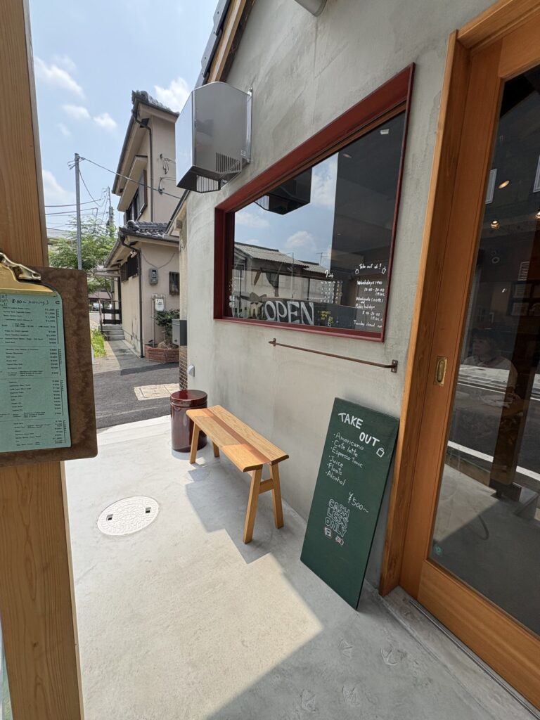 鴨葱喫茶の入口。コンクリートの土間に木のベンチ、赤い枠の窓に OPEN の文字と営業時間が手書きされている