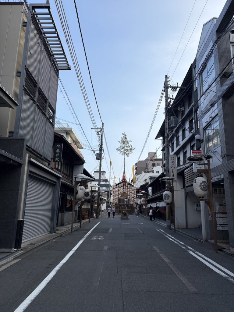 朝の山鉾町の通り。奥に白い提灯を掲げた山鉾が立ち、真木の松が空へ伸びている