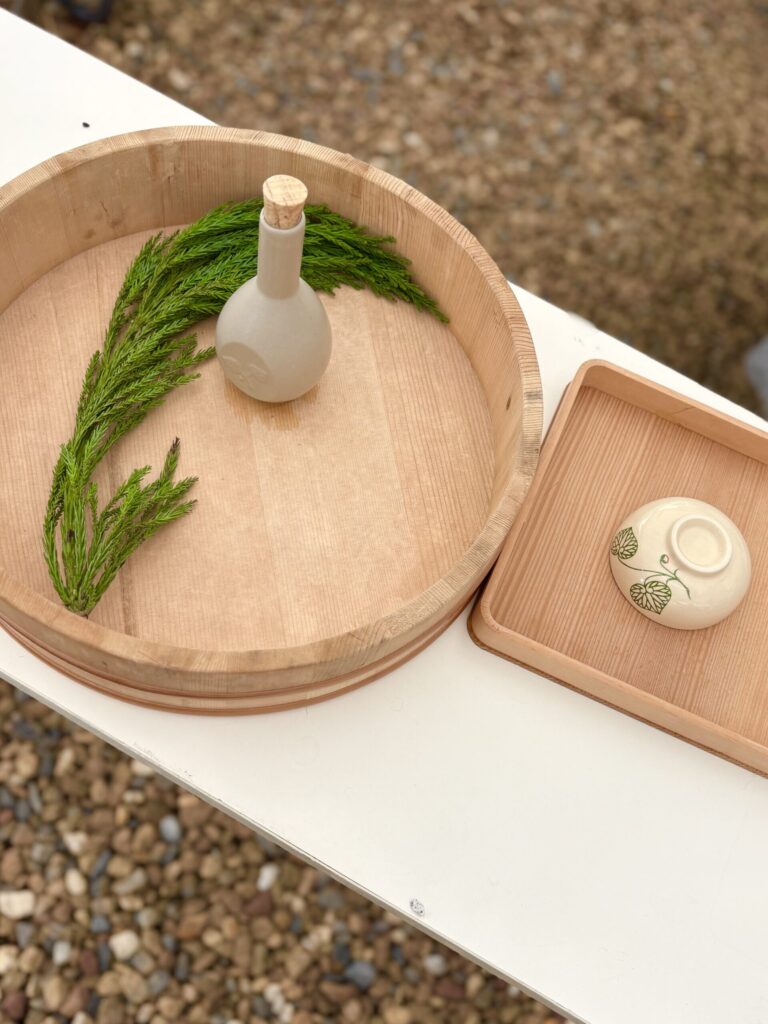 白木の丸桶に置かれた白い徳利と杉の葉、隣の角盆には草花模様の白い小皿