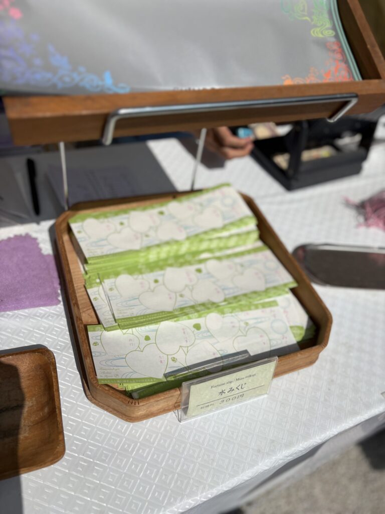 水みくじの授与所。木のトレイに緑の水みくじが積まれ、初穂料三百円の札が立つ