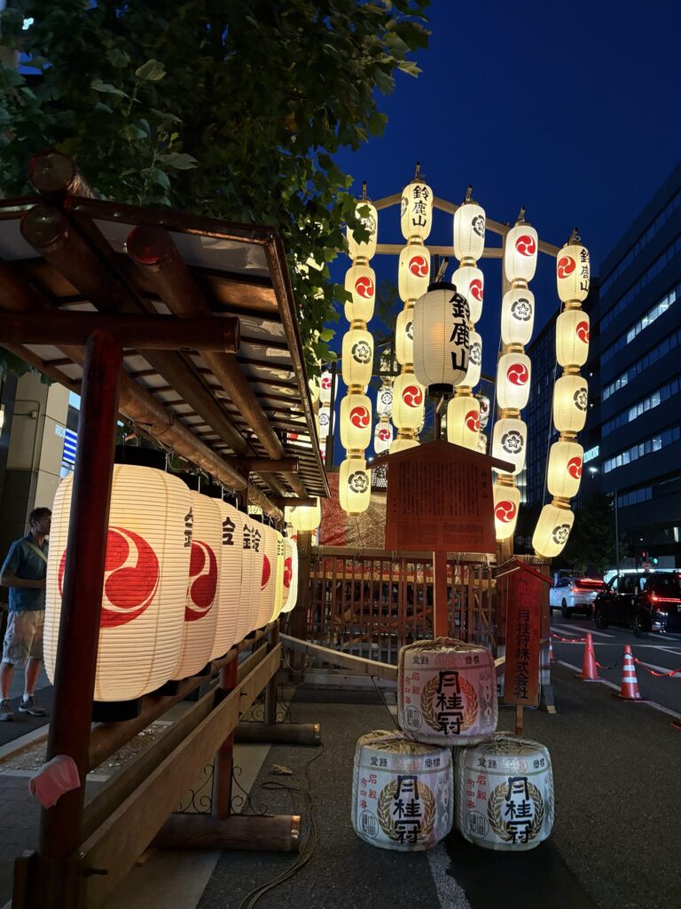 祇園祭 後祭・鈴鹿山の駒形提灯と駒札。奉献の酒樽が積まれ、暮れかけた空に灯りがともる