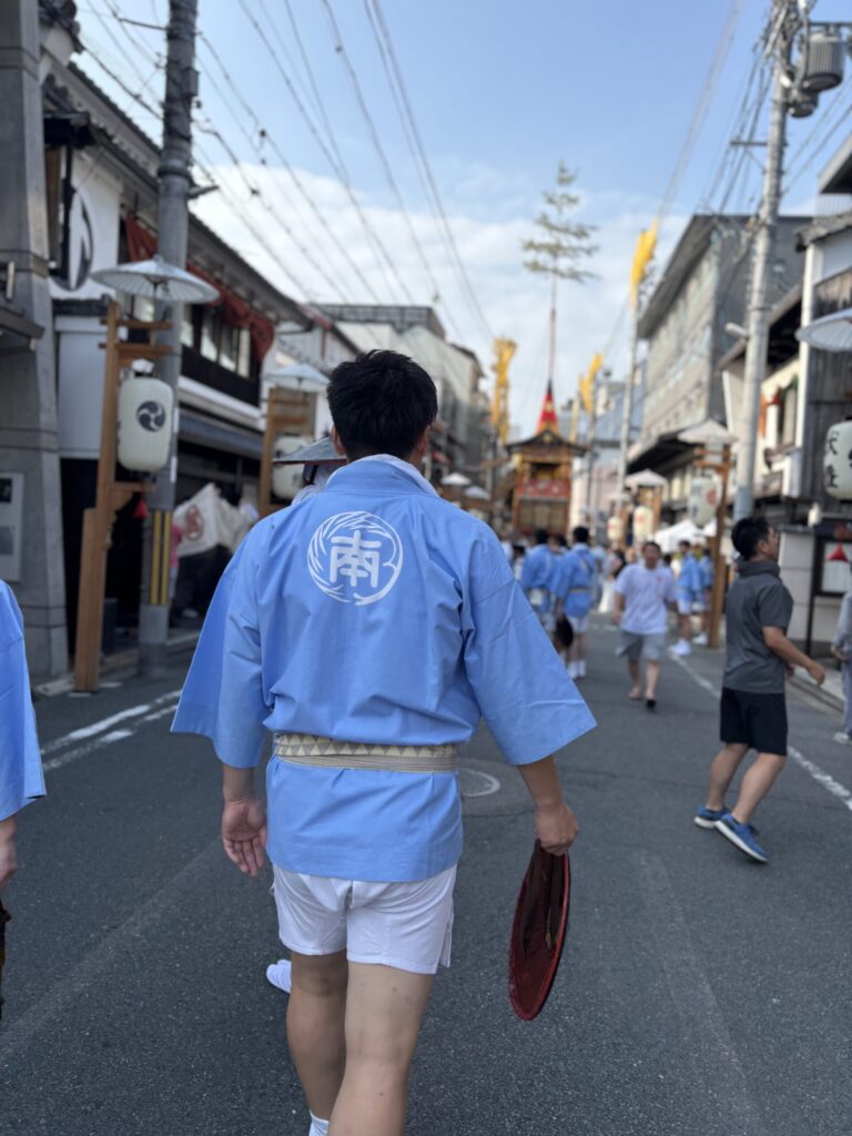 祇園祭 後祭・巡行の朝。「南」の法被姿の人と、通りの奥に立つ真松