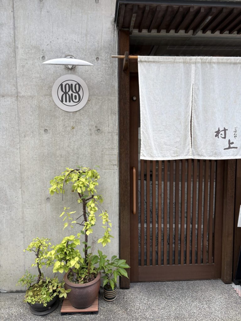 ごはんや村上の外観。コンクリートの壁に丸いロゴ、白い暖簾と鉢植え
