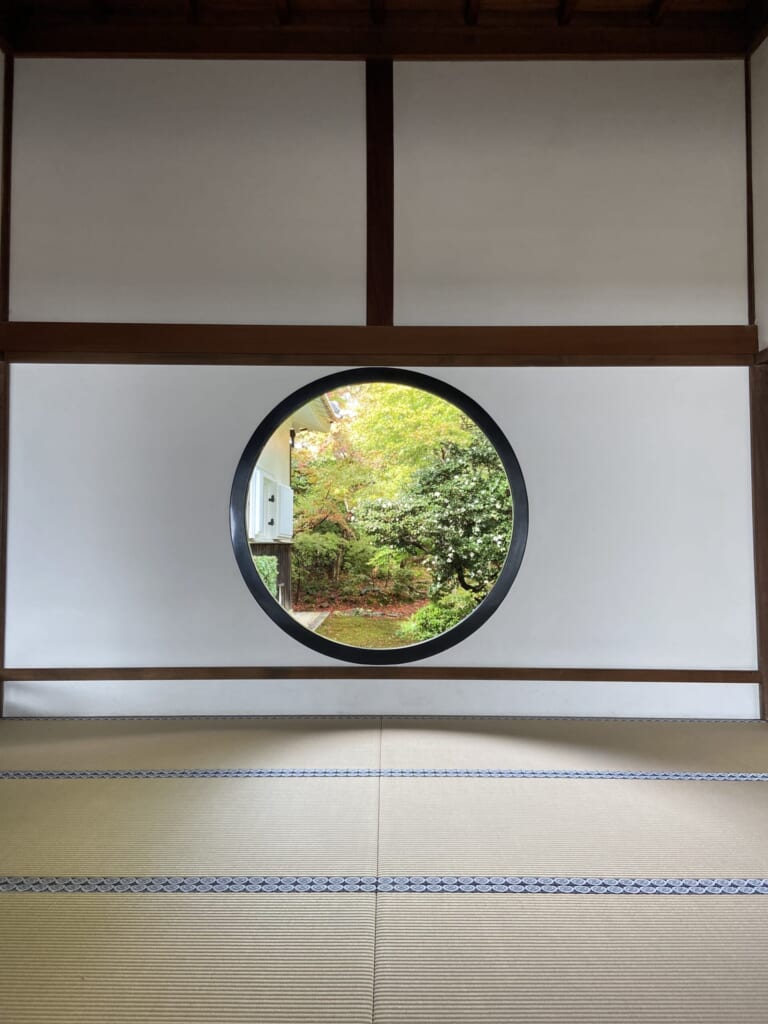 源光庵「悟りの窓」。白い壁の丸窓の向こうに、もみじと白い花の咲く庭が見える